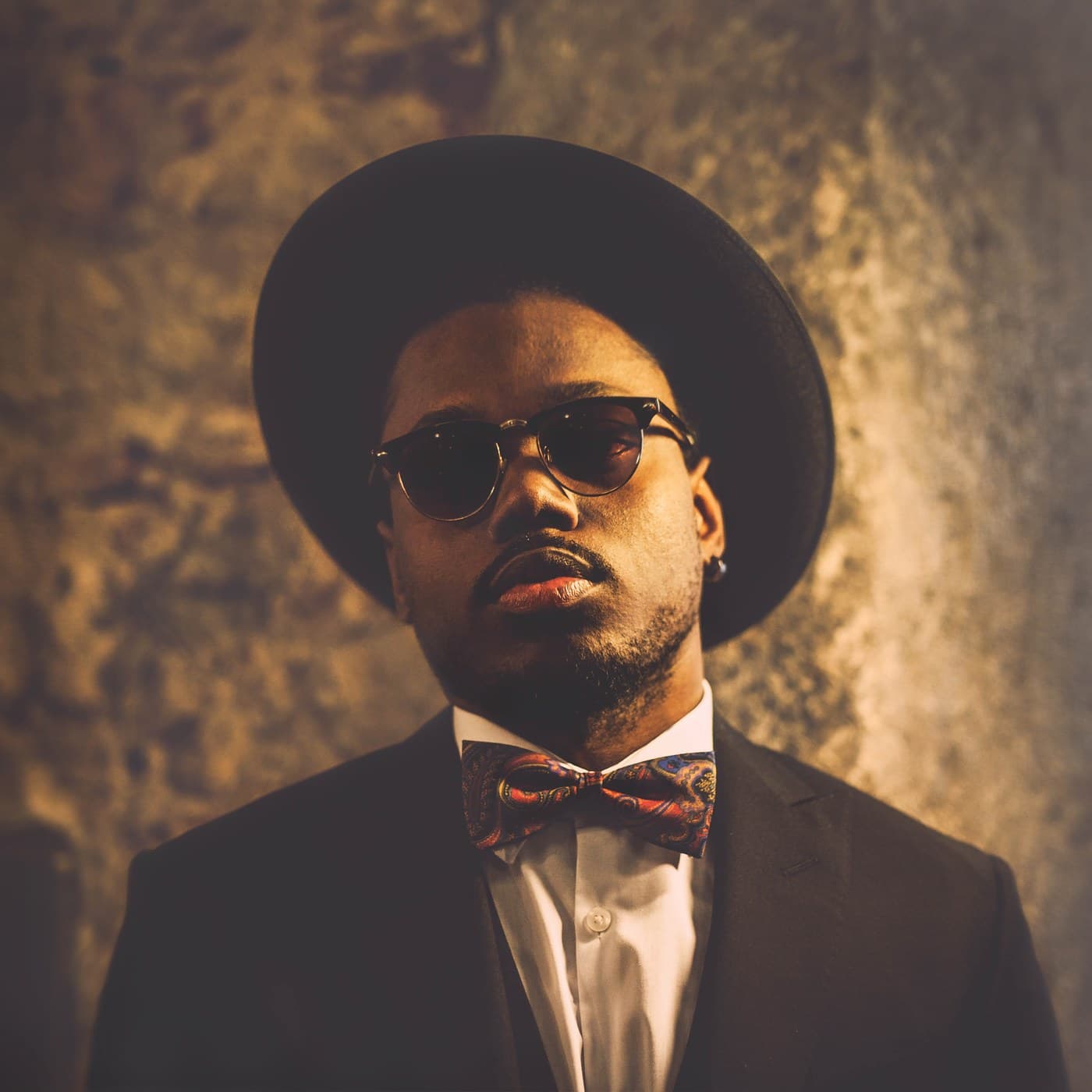 Editorial portrait — wide-brim hat, round sunglasses, bow tie, warm stone wall