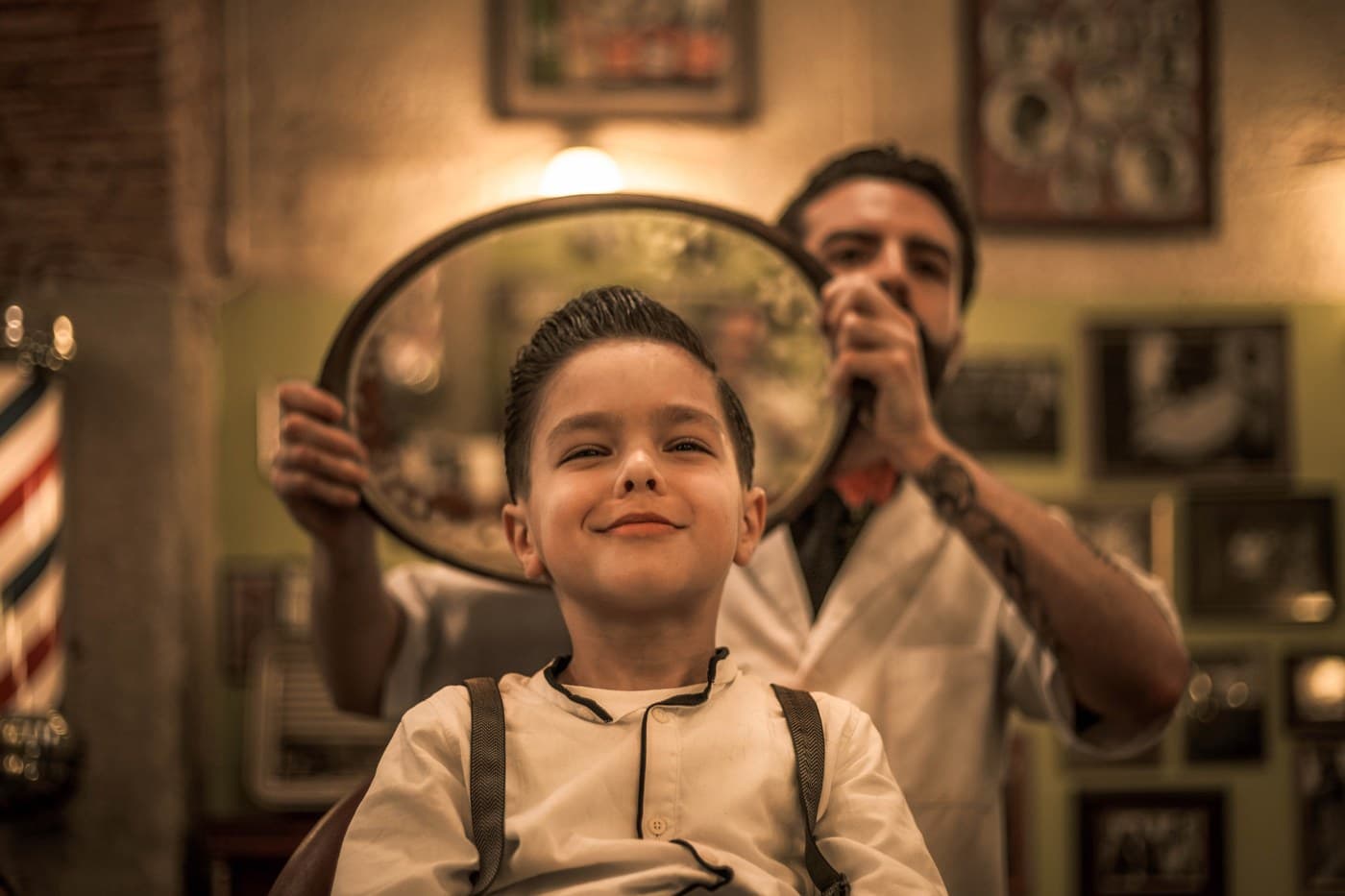 Happy young boy after his first haircut, barber reveals the result with oval mirror