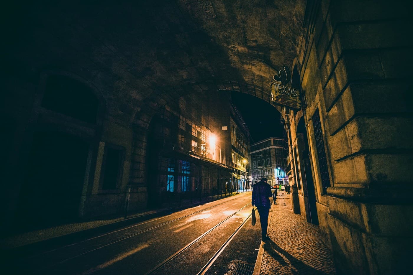 Cinematic night scene in Lisbon — figure under arch with TEXAS neon sign, tram tracks