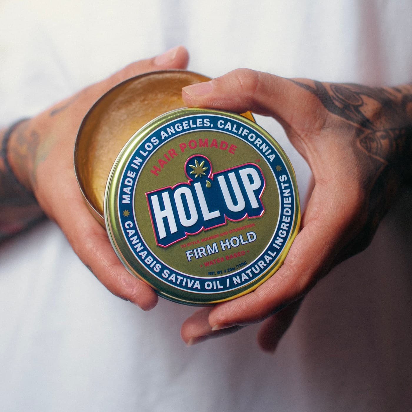 HOL'UP hair pomade tin held open by tattooed hands, soft studio light