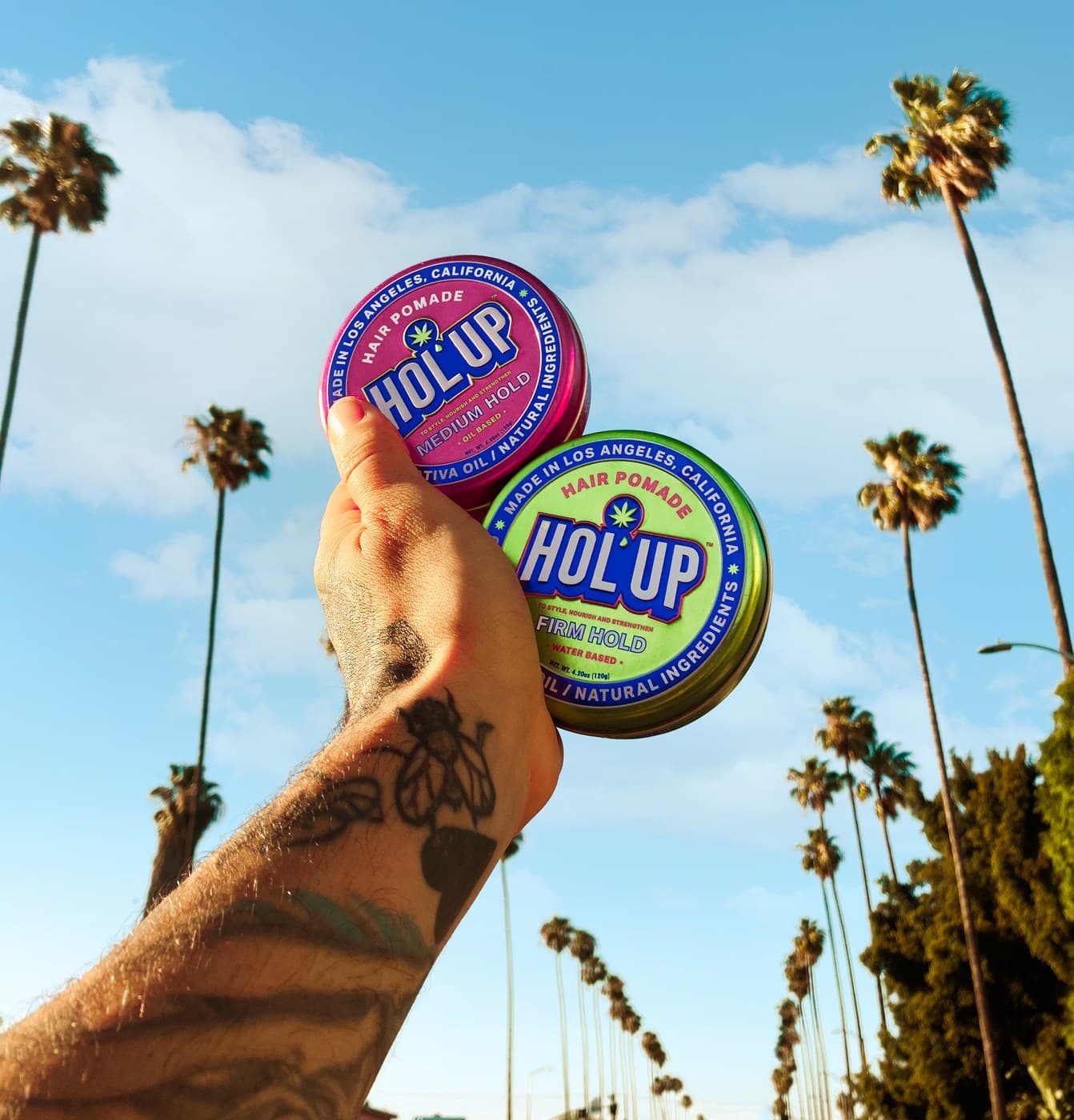 Tattooed hand holding HOL'UP pomade tins up against LA blue sky and palm trees